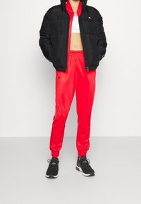 Giacca a vento nera con chiusura a zip, pantaloni sportivi rossi e giacca a vento nera. Il materiale appare trapuntato, liscio e lucido.