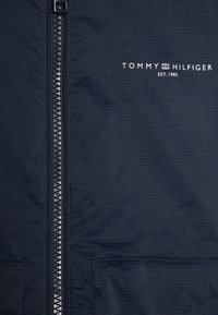 Mornarsko modra jakna z dolgim zadrgo, teksturirano blago in belim logotipom "TOMMY HILFIGER EST. 1985" na zgornji levi strani.