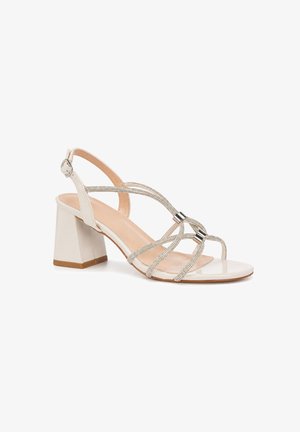 Sandale à talon bloc beige unique avec fines lanières métalliques croisées sur le pied et fermeture à boucle à la cheville.