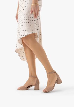 Nogi w beżowych sandałkach na słupku z paskiem na kostce, połączone z białą spódnicą o asymetrycznym brzegu w czarne grochy oraz złotą biżuterią na ręce.