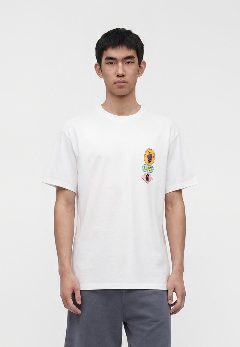 Junger Mann, der ein weißes T-Shirt mit einer kleinen bunten Grafik von Trauben und Formen auf der linken Brust trägt, kombiniert mit grauen Shorts.