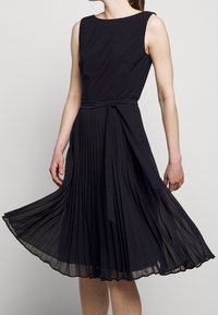 Žena v černých skládacích šatech bez rukávů s opaskem uvázaným v pase, drží sukni mírně zvednutou proti jednobarevnému pozadí.