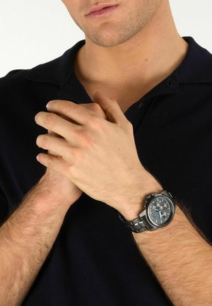 Uomo con camicia nera che stringe le mani vicino al petto, indossando un orologio da polso in metallo nero con quadrante con accenti blu al polso sinistro.