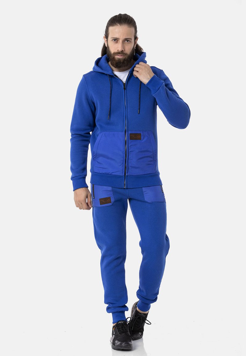 Blauer Sweatshirt mit Reißverschluss und Kapuze, ausgestattet mit Vordertaschen. Kombiniert mit blauen Hosen, beide aus weichem Stoff mit regulärer Passform.