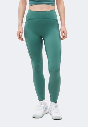 Nő, aki türkizkék magasderekú leggingset és fehér sportcipőt visel, egyszínű háttér előtt áll.