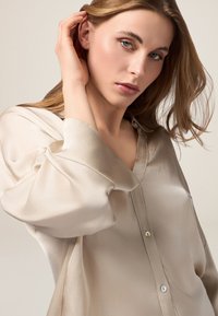Blusa in satin beige chiaro con una vestibilità rilassata, caratterizzata da un patte, maniche morbide drappeggiate e una leggera lucentezza. Texture liscia, bottoni semplici.