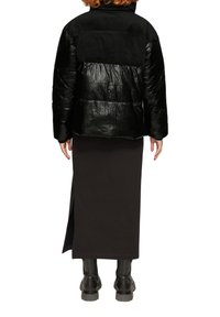 Zwarte pufferjack met gekreukelde textuur en corduroy accenten, gecombineerd met een lange zwarte rok en zwart laarzen tot de knie.