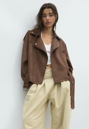 Jeune femme portant une veste marron oversize, un crop top blanc et un pantalon beige large, debout sur un fond clair uni.