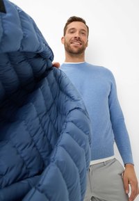 Dunkelblauer Pufferjacke mit gestepptem Muster, teilweise geöffnet, sodass das weiche Innenfutter sichtbar ist. Das Modell trägt einen hellblauen Pullover und graue Hosen.
