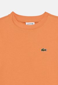 Πορτοκαλί βαμβακερό T-shirt με στρογγυλή λαιμόκοψη, που φέρει μικρό πράσινο και λευκό κεντητό λογότυπο κροκόδειλου στο στήθος.