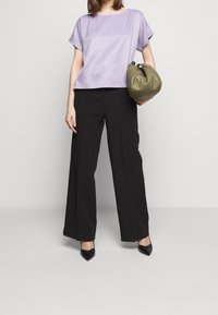Donna che indossa una maglietta lavanda a maniche corte, pantaloni neri a gamba larga, tacchi neri a punta, mentre tiene una borsa verde oliva all'altezza della vita.