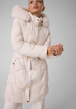 Un mannequin porte un long manteau matelassé couleur crème, bordé de fourrure moelleuse autour de la capuche et orné de boutons dorés. Elle tient les mains jointes devant elle, sur un fond gris uni.