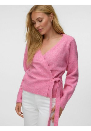 Femme portant un pull enveloppant rose duveteux noué à la taille et un pantalon blanc, regardant vers le bas, les cheveux blonds tombant sur une épaule.