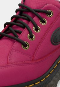 Chaussure Dr. Martens en cuir rose vif avec lacets noirs, œillets dorés, semelle noire et détail de couture jaune.