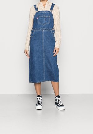 Kvinde iført en blå denim midi overalls kjole over en beige ternet langærmet skjorte, kombineret med sort-hvide høje sneakers.