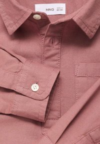 Růžová košile s krátkým rukávem vyrobená z texturované tkaniny, vybavená límcem na knoflíčky, přední kapsou a standardním zapínáním na knoflíky.