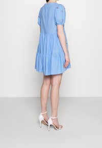 Mujer que lleva un vestido corto de mangas cortas en azul con bordados florales y sandalias de tacón alto blancas con tira en el tobillo, vista por detrás.