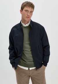 Marine bomberjack met ritssluiting, ribbels aan de manchetten en twee zijzakken. Gelaagd over een groene trui en een witte blouse, gecombineerd met beige broeken.