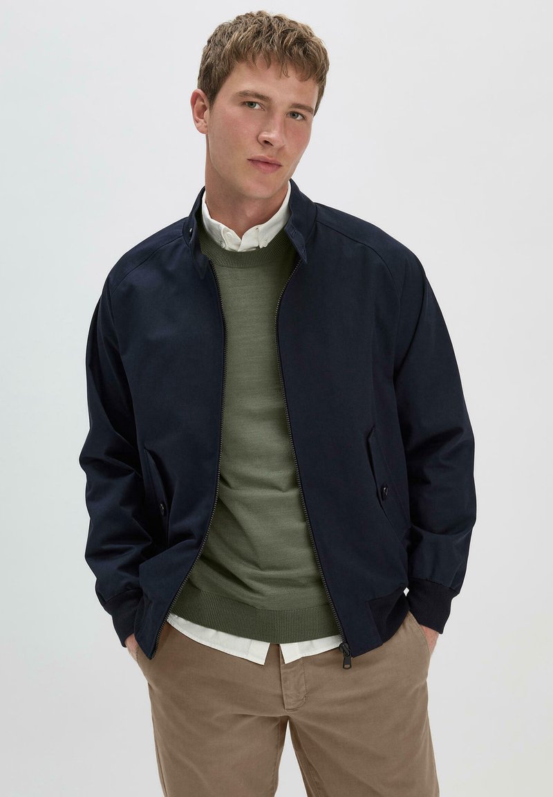 Marine bomberjack met ritssluiting, ribbels aan de manchetten en twee zijzakken. Gelaagd over een groene trui en een witte blouse, gecombineerd met beige broeken.