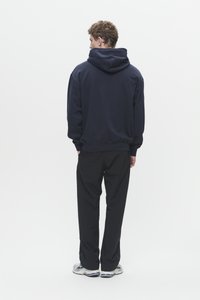 Un uomo dai capelli ricci sta in piedi di spalle, indossando un hoodie blu navy, pantaloni neri e sneakers bianche con dettagli blu.