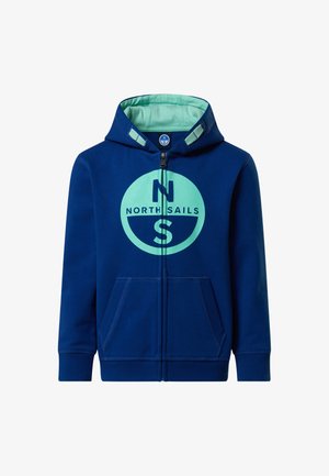 Garçon debout, une main dans la poche, portant un sweat à capuche zippé bleu North Sails, un pantalon bleu marine et des baskets blanches à scratch sur un fond uni.