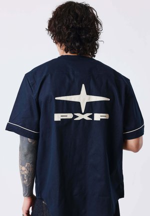 Hombre con cabello oscuro y rizado y brazo tatuado que lleva una camisa azul marino con una estrella blanca y el texto "PXP" en la espalda, de pie contra un fondo claro y sencillo.