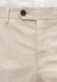 Μπεζ καρό παντελόνι στερεωμένο με σκούρο καφέ κουμπί, σε συνδυασμό με λευκό πουκάμισο μπαλωμένο στη μέση της μέσης.