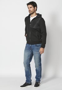 Felpa nera con zip, texture a coste e pannello lucido, abbinata a jeans in denim blu e sneakers nere. Cappuccio con lacci regolabili.