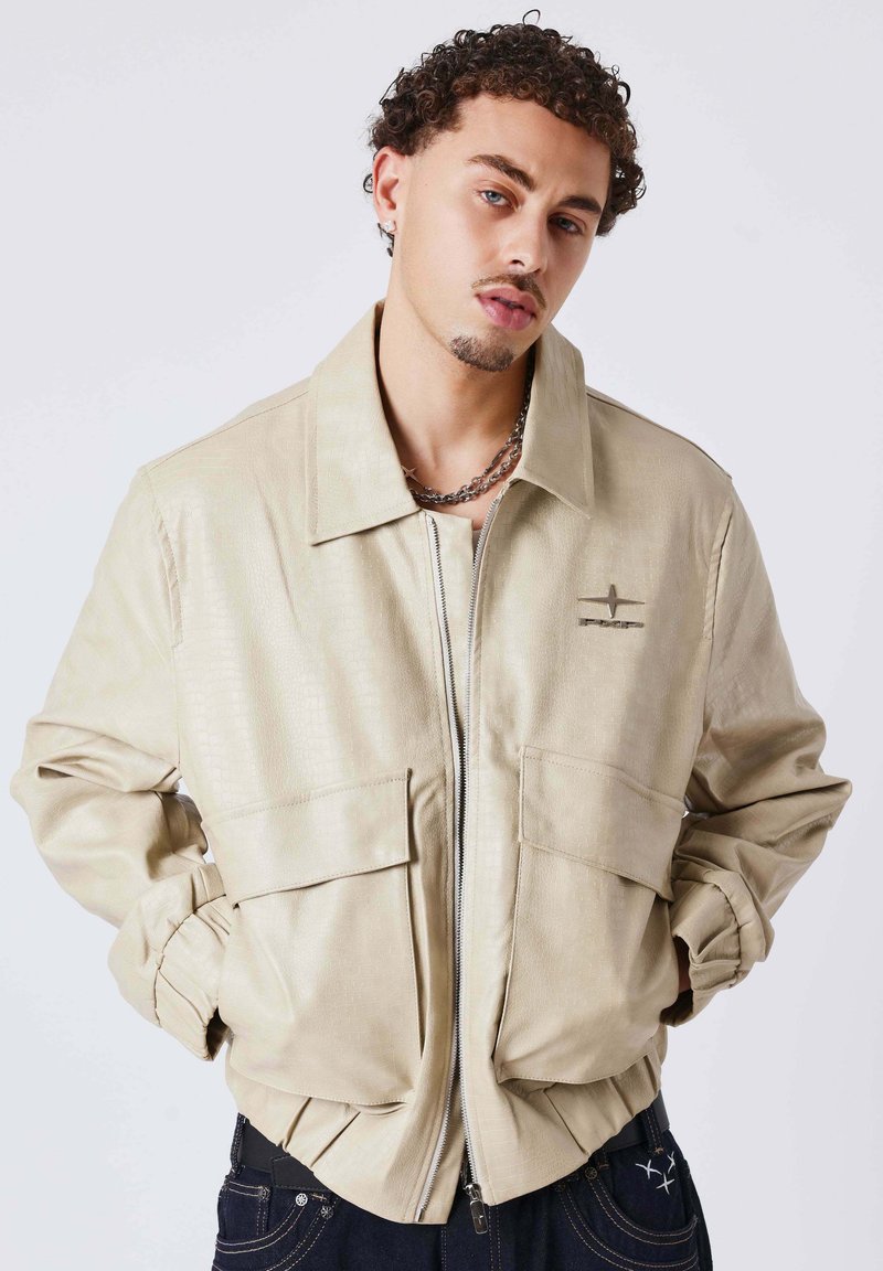 Beige jas met krokodiltextuur, voorzien van een kraag, rits aan de voorkant en twee grote klepzakken. Heeft gerimpelde manchetten en een klein logodetail.