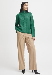 Grüner Kabelstrickpullover mit hohem Kragen und Reißverschlussdetail, kombiniert mit beigen weit geschnittenen Hosen und schwarzen spitzen High Heels.