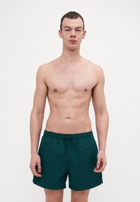 Shorts de bain verts avec une taille élastique et un cordon de serrage. Fabriqués en matériau texturé, avec des fentes latérales pour plus de facilité de mouvement.