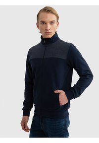 Jonge man met blond haar, gekleed in een marineblauw en donkergrijs jack met rits en blauwe jeans, staand met één hand in zijn zak.