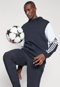 Haut de survêtement noir et blanc avec poche kangourou, associé à un pantalon noir. Un ballon de football est équilibré sur le genou du mannequin.