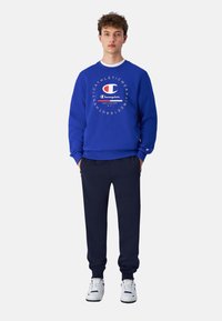 Blauwe sweatshirt met een rond logo op de borst, gecombineerd met donkerblauwe joggingsbroek en witte sportschoenen. Glad materiaal, comfortabele pasvorm.