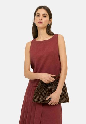 Mujer con vestido sin mangas de color rojo oscuro que sostiene un bolso de mano marrón con textura de cuentas sobre un fondo liso.