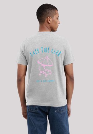 Man wearing gray t-shirt with blue text "Lazy Tide Club" and pink beach chair with umbrella graphic on the back, paired with blue jeans.