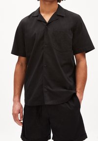 Czarna koszula z krótkim rękawem wykonana z lekkiego materiału, z zapinanym na guziki przodem, kieszenią na piersi i luźnym kołnierzem.