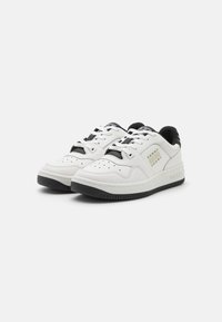 Zapatillas blancas de cuero con suela de goma negra, que presentan detalles en negro, perforaciones y un logo en el costado.