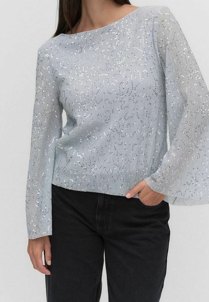 Femme portant un pull argenté à manches longues avec des paillettes brillantes et un pantalon noir, la main partiellement dans la poche, sur un fond uni.