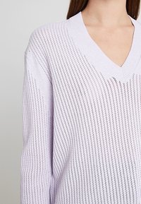 Femme portant un pull violet clair à col en V avec un motif rayé en maille ajourée verticale, visible des épaules jusqu'au milieu du torse.