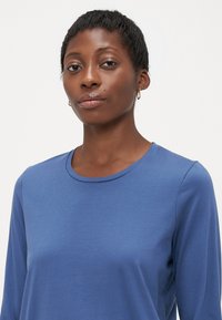 Kvinna med kort hår som bär guldörhängen och en blå långärmad skjorta, vänd framåt mot en enfärgad ljus bakgrund.