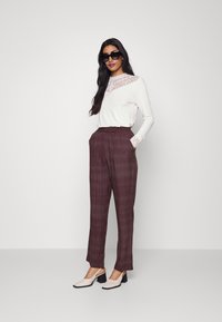 Blusa branca de manga longa com detalhes em renda, combinada com calças padrão em cor bordô. Modelo usa óculos de sol pretos e sapatos de salto bloco bege.