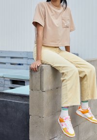 Person i beige kortärmad skjorta, gula kordflöjtsbyxor, vita strumpor med gröna ränder och vita, orange och rosa Nike-sneakers som sitter på betongblock.