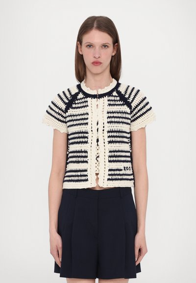 Hæklet cardigan med korte ærmer, marineblå og cremefarvede striber, scalloped halsudskæring, knapper foran og en dekorativ kant. Matchet med mørke shorts.