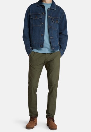 Veste en denim bleu foncé avec poches poitrine, t-shirt bleu clair, pantalon vert olive et bottes marron à lacets. Ensemble décontracté.