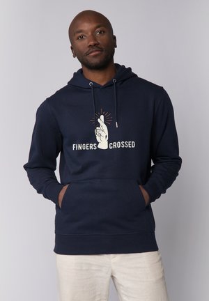 Man draagt een marineblauwe hoodie met een graphic van gekruiste vingers en de tekst "FINGERS CROSSED", handen in de voorkantzak, beige broek.