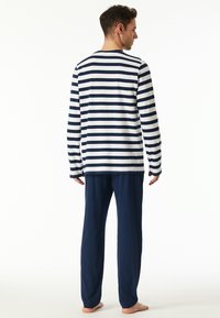 Mann, der barfuß steht und wegschaut, trägt ein langärmliges, weiß-blau gestreiftes Hemd und einfarbige marineblaue Hose vor weißem Hintergrund.