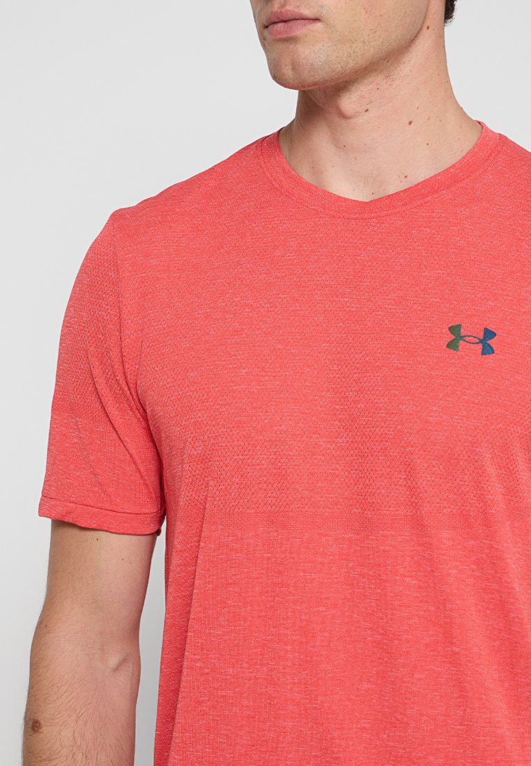 Mężczyzna w koralowo-czerwonej koszulce sportowej Under Armour z krótkim rękawem, z logo na lewej piersi, na jednolitym tle.