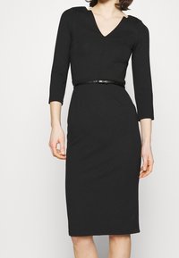 Robe noire ajustée avec un décolleté en V, des manches trois-quarts et une fine ceinture noire. Fabriquée en tissu lisse, conception à mi-genoux.