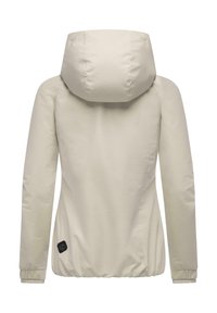 Beżowa kurtka z kapturem, z długimi rękawami, elastycznymi mankietami oraz elastycznym dołem, z małą czarną naszywką na dolnym lewym boku.